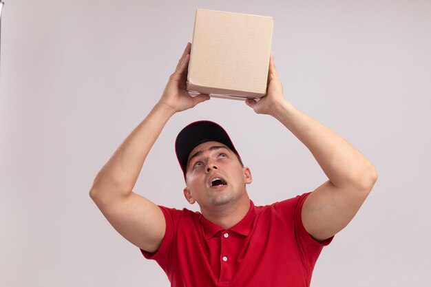 Thinking young delivery man wearing uniform with cap raising and looking at box isolated on white wall