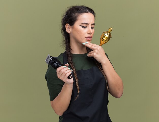 Думающая молодая женщина-парикмахер в униформе держит машинку для стрижки волос с кубком победителя, положив пальцы на подбородок, изолированные на оливково-зеленой стене