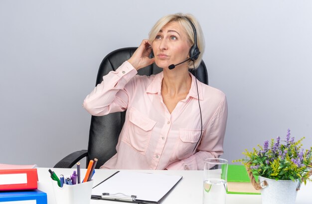 думающая молодая женщина-оператор call-центра в гарнитуре сидит за столом с офисными инструментами, изолированными на белой стене