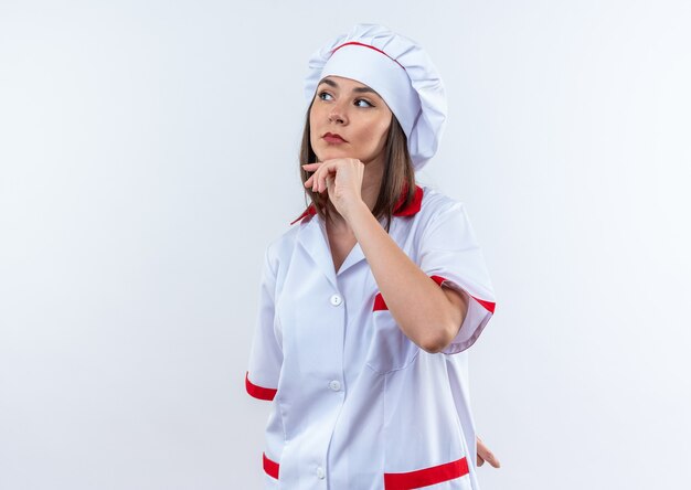 думающая молодая женщина-повар в униформе шеф-повара схватилась за подбородок, изолированную на белой стене