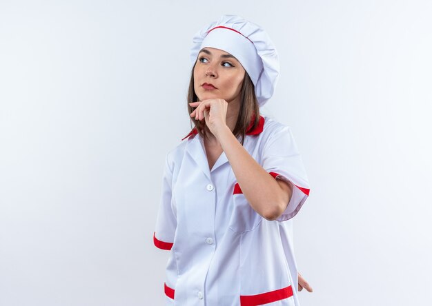 thinking young female cook wearing chef uniform grabbed chin isolated on white wall