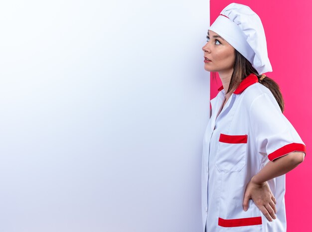 Thinking young female cook wearing chef uniform stands nearby white wall isolated on pink background with copy space