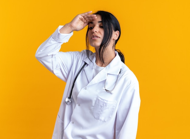 Foto gratuita pensando giovane dottoressa indossando accappatoio medico con stetoscopio guardando a distanza con la mano isolata su sfondo giallo