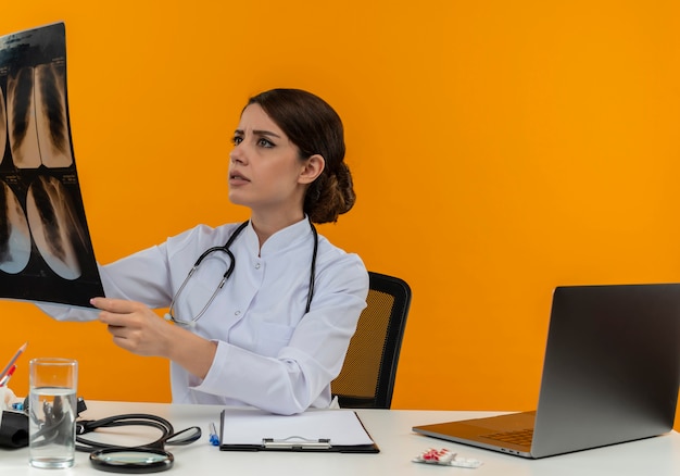 Thinking young female doctor wearing medical robe with stethoscope sitting at desk work on computer with medical tools holding and looking at x-ray on isolation yellow background