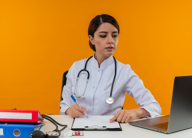 Думающая молодая женщина-врач в медицинском халате со стетоскопом, сидя за столом, работает на компьютере с медицинскими инструментами, глядя на ноутбук на изолированном желтом фоне