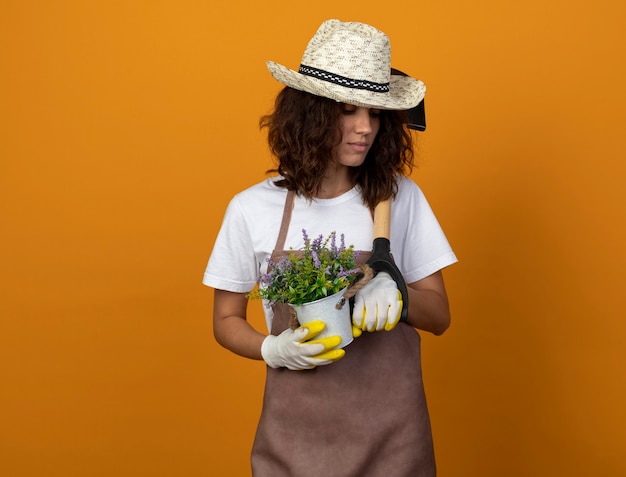 Thinking young female gardener in uniform wearing gardening hat and gloves holding flower in flowerpot and putting spade on shoulder