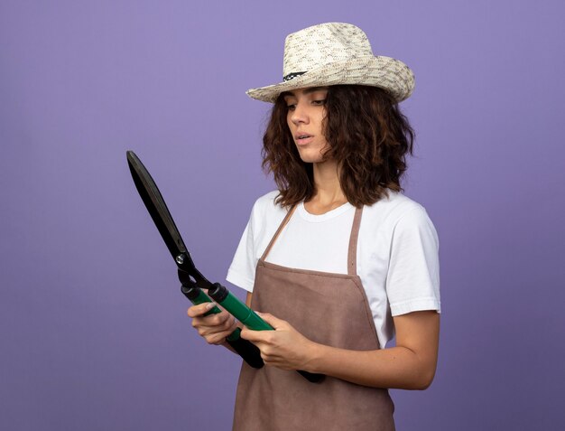 Thinking young female gardener in uniform wearing gardening hat holding and looking at clippers