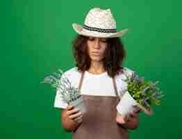 Foto gratuita pensando giovane giardiniere femminile in uniforme che indossa cappello da giardinaggio tenendo e guardando i fiori in vasi da fiori