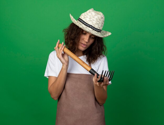 Free photo thinking young female gardener in uniform wearing gardening hat holding and looking at rake