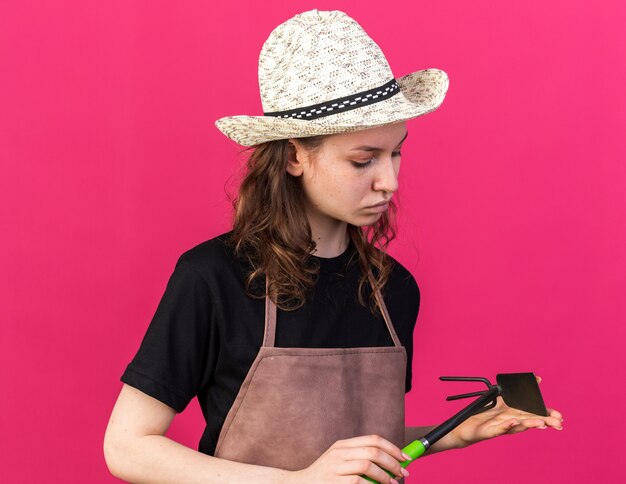 Thinking young female gardener wearing gardening hat holding and looking at hoe rake isolated on pink wall