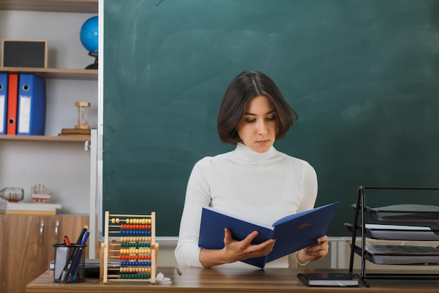 thinking young female teacher holding and reading book sitting at desk with school tools on in classroom