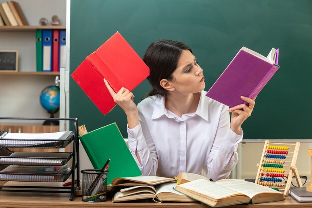 Thinking young female teacher holding and reading book sitting at table with school tools in classroom