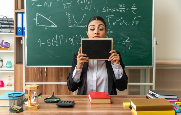 思考の若い女性教師は、教室でミニ黒板を持って見ている学用品と一緒にテーブルに座っています