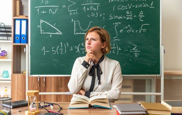 생각하는 젊은 여성 교사가 교실에서 턱을 잡은 학교 도구로 테이블에 앉아있다.