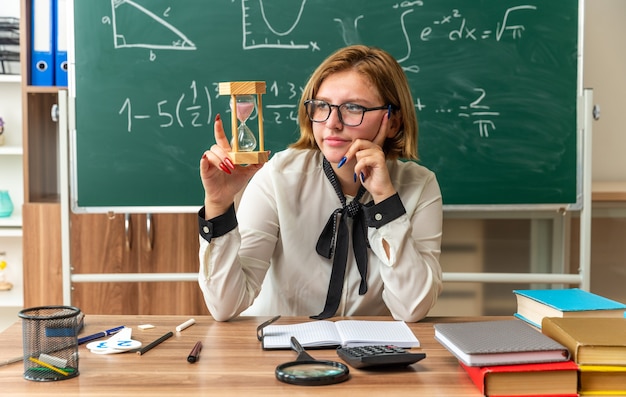 Thinking young female teacher sits at table with school tools holkding and looking at hourglass in classroom