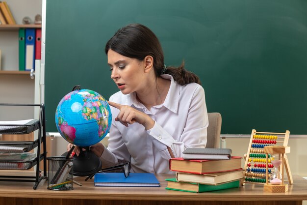 Thinking young female teacher sitting at table with school tools holding and putting finger on globe in classroom