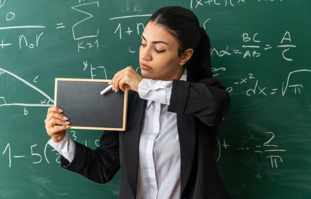 Thinking young female teacher standing in front blackboard holding mini blackboard with stranded for board in classroom