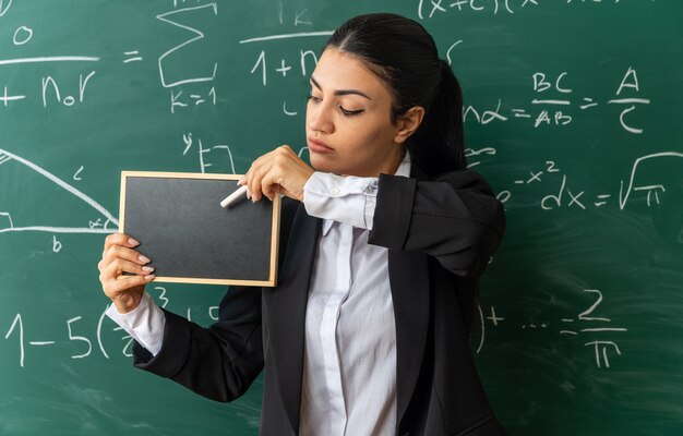 教室でボードのために座礁したミニ黒板を保持している前の黒板に立っている若い女教師を考える