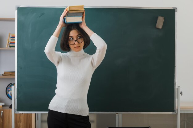 thinking young female teacher wearing glasses holding books on head standing in front blackboard in classroom