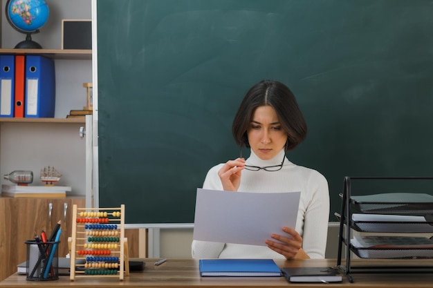 thinking young female teacher wearing glasses holding and looking at paper sitting at desk with school tools on in classroom