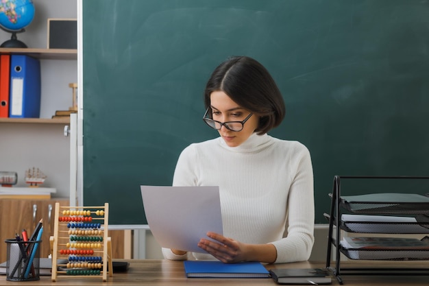 眼鏡をかけている若い女教師を考えて、教室で学校の道具を持って机に座っている紙を見て