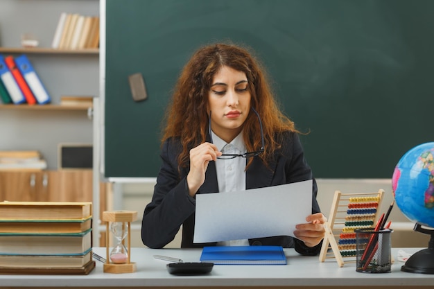 眼鏡をかけて、教室で学校の道具を持って机に座って紙を読んでいる若い女性教師を考える