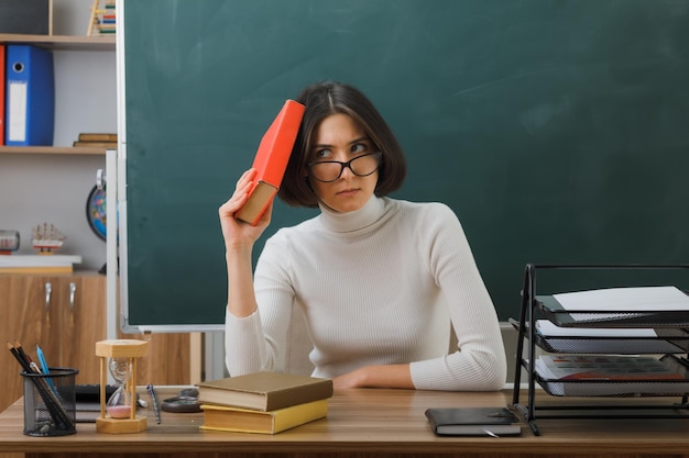 교실에서 학교 도구를 사용하여 책상에 앉아 책을 머리에 올려놓는 안경을 쓴 젊은 여교사