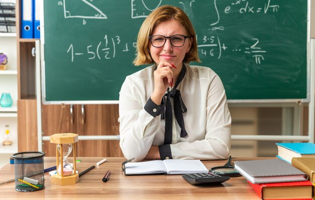 thinking young female teacher wearing glasses sits at table with school supplies grabbed chin in classroom