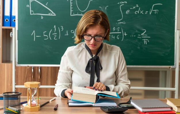 Thinking young female teacher wearing glasses sits at table with school tools reading book in classroom