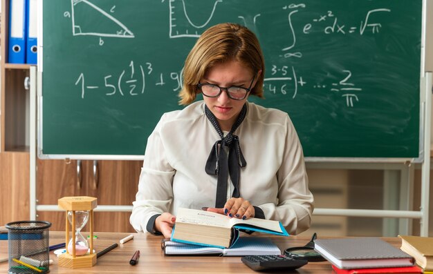 眼鏡をかけている思考の若い女性教師は、教室で本を読んで学校のツールとテーブルに座っています