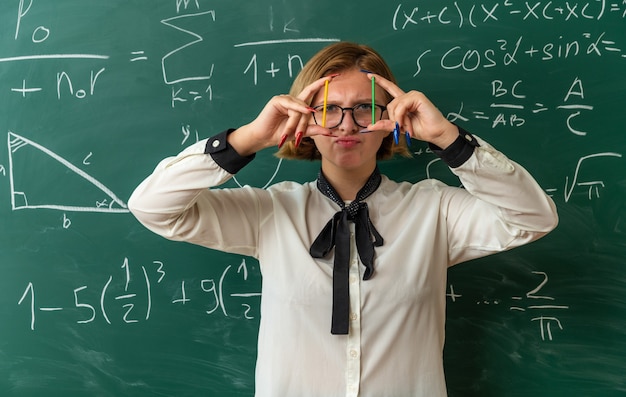 thinking young female teacher wearing glasses standing in front blackboard holding pencils in classroom