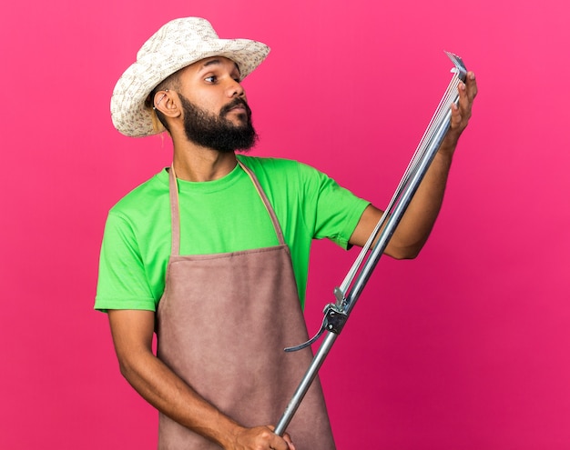 Thinking young gardener afro-american guy wearing gardening hat holding and looking at leaf rake 