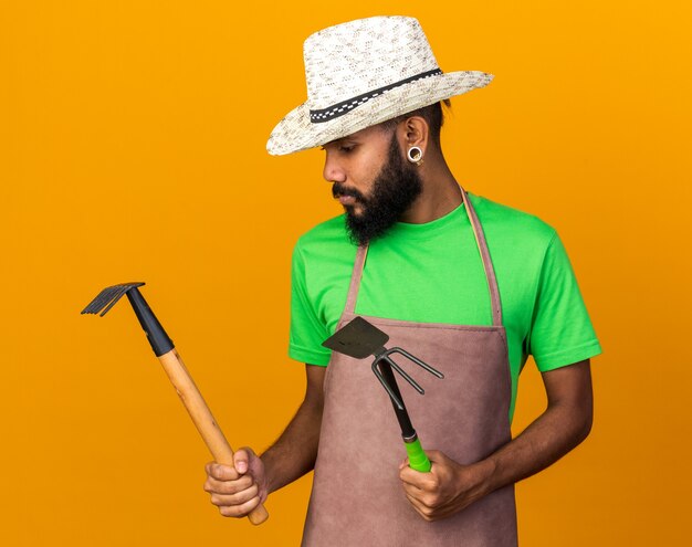 Thinking young gardener afro-american guy wearing gardening hat holding and looking at rake with hoe rake 