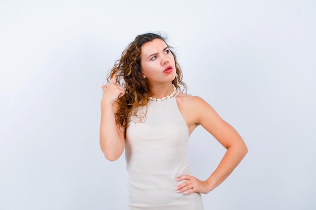 Thinking young girl is holding hair and putting other hand on waist on white background