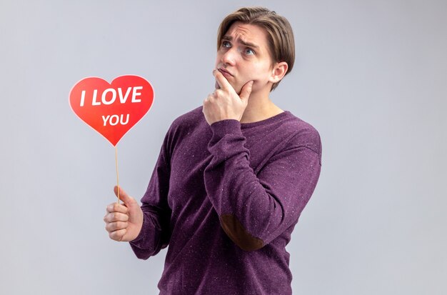 Thinking young guy on valentines day holding red heart on a stick with i love you text putting hand on chin isolated on white background