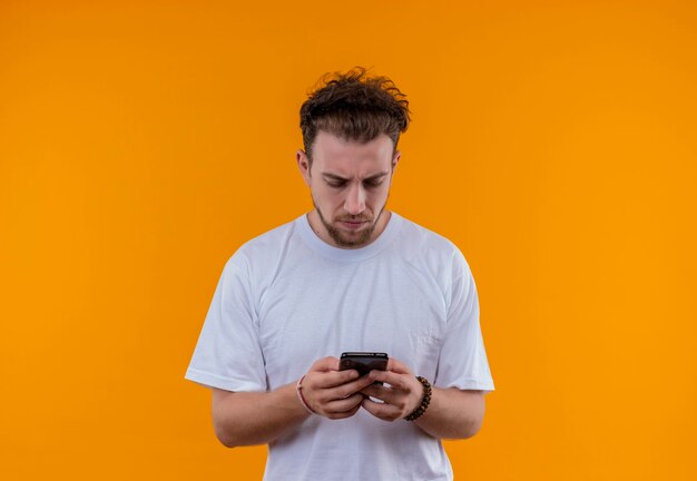孤立したオレンジ色の壁に手元の携帯電話を見て白いTシャツを着て考える若い男
