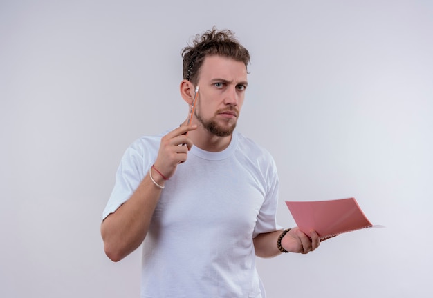 Thinking young guy wearing white t-shirt put pencil on cheek holding notebook on isolated white wall