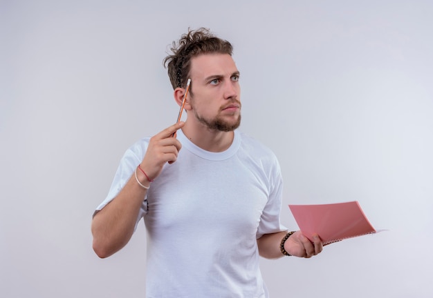 Thinking young guy wearing white t-shirt put pencil on forehead holding notebook on isolated white wall