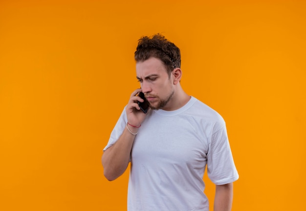 흰색 티셔츠를 입고 생각하는 젊은 남자가 고립 된 주황색 벽에 전화로 말한다.