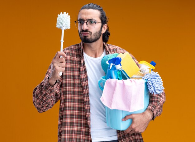 Thinking young handsome cleaning guy wearing t-shirt holding bucket of cleaning tools and looking at brush in his hand isolated on orange wall