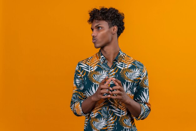 Thinking young handsome dark-skinned man with curly hair in leaves printed shirt holding hands together on an orange background