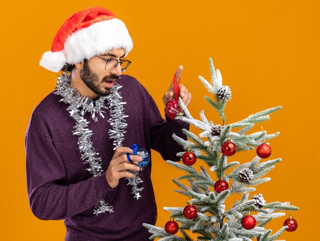 Thinking young handsome guy standing nearby christmas tree wearing christmas hat with garland on neck holding christmas balls isolated on orange background