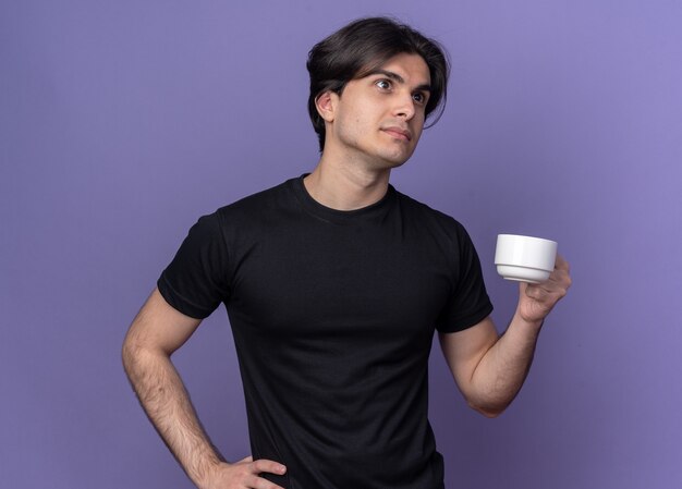 Думающий молодой красивый парень в черной футболке держит чашку кофе, положив руку на бедро, изолированную на фиолетовой стене