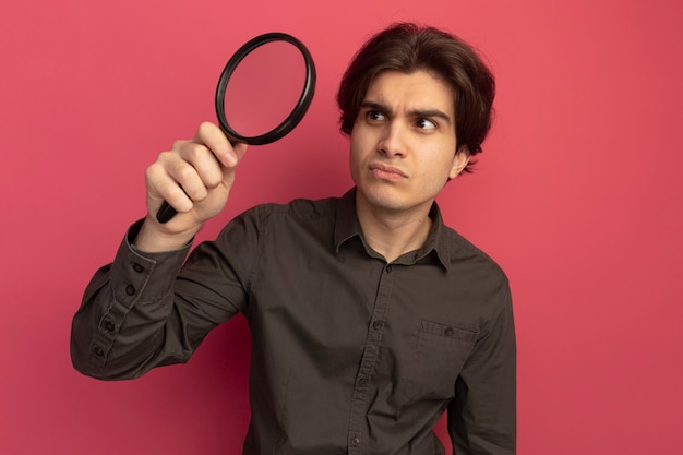 Thinking young handsome guy wearing black t-shirt holding and looking at magnifier isolated on pink wall