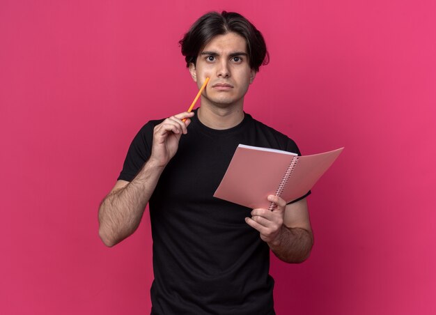 Думающий молодой красивый парень в черной футболке держит блокнот с карандашом на розовой стене