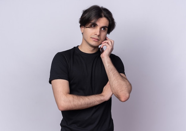 Thinking young handsome guy wearing black t-shirt speaks on phone isolated on white wall