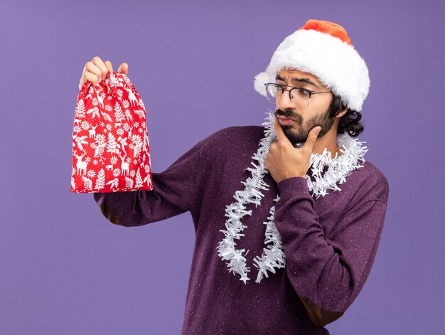 Thinking young handsome guy wearing christmas hat with garland on neck holding and looking at christmas bag isolated on blue background