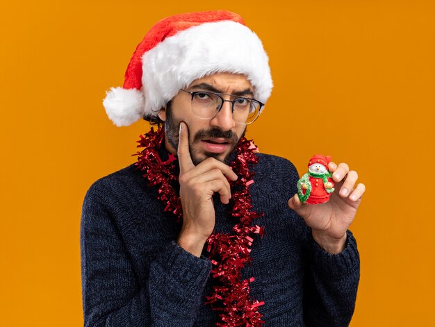 Thinking young handsome guy wearing christmas hat with garland on neck holding toy putting finger on cheek isolated on orange background