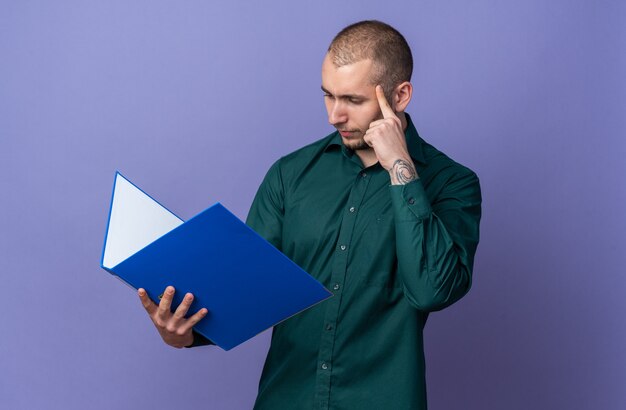 Thinking young handsome guy wearing green shirt holding and looking at folder 