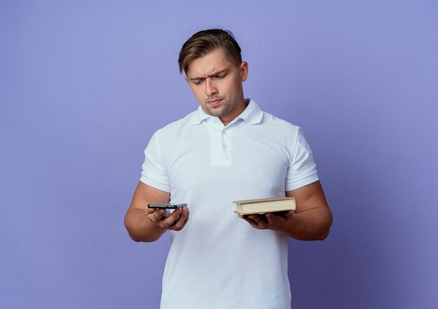 Thinking young handsome male student holding and looking at phone with book isolated on blue